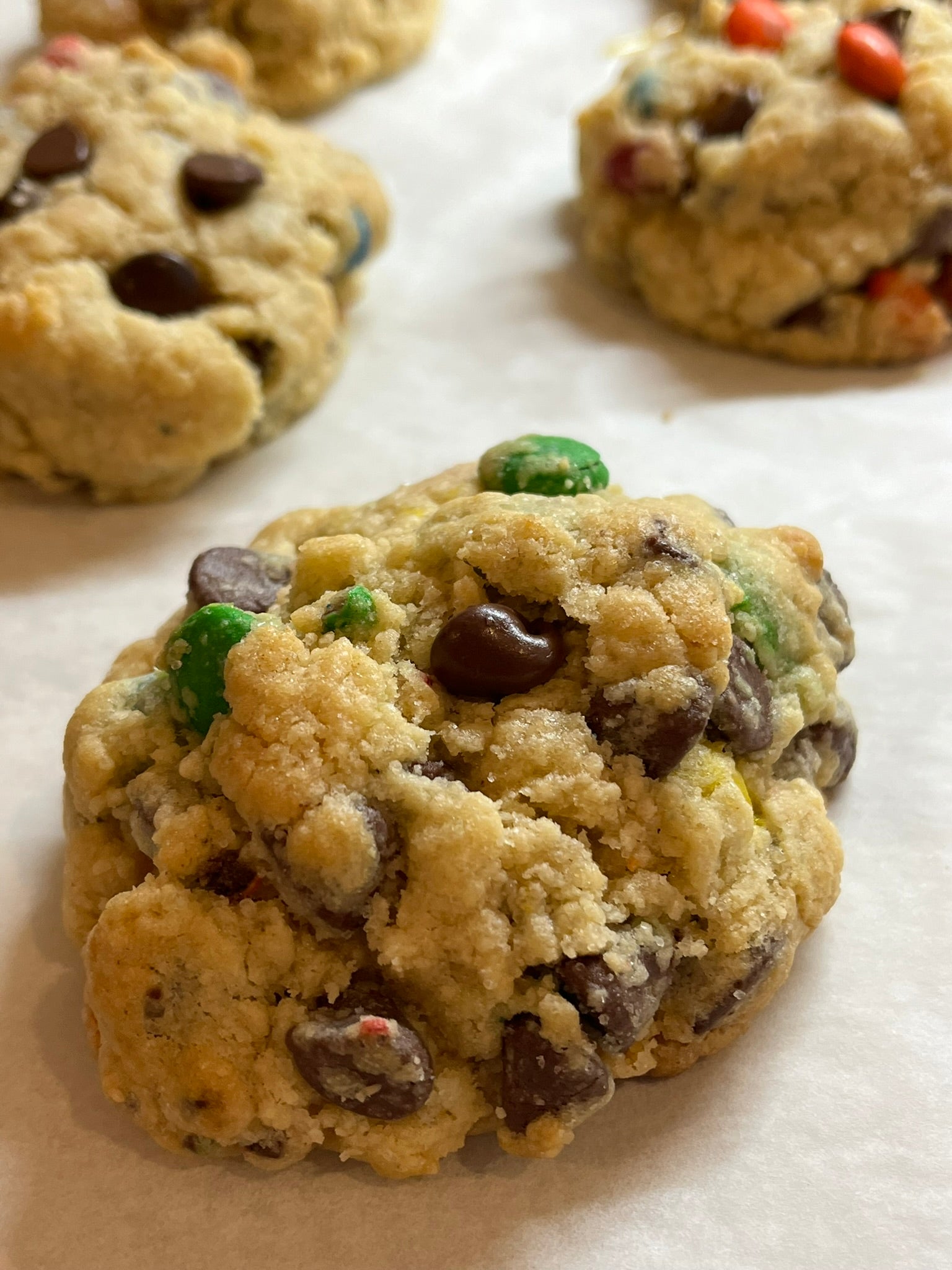 A close-up of a golden-brown chocolate chip cookie with green and red candy pieces, placed on a white surface with other cookies blurred in the background.